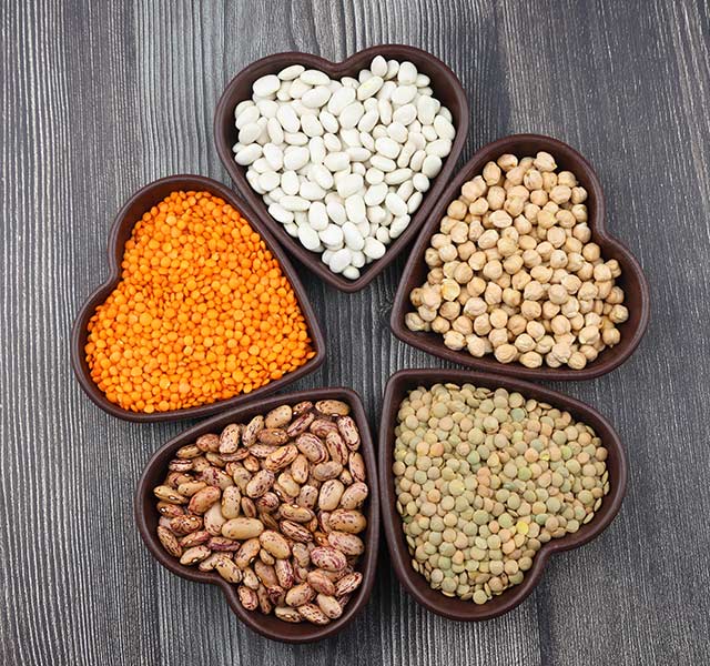 Heart-shaped bowls contain a variety of legumes on a dark wooden surface. The bowls hold white beans, red lentils, chickpeas, pinto beans, and green lentils. beans for blood pressure