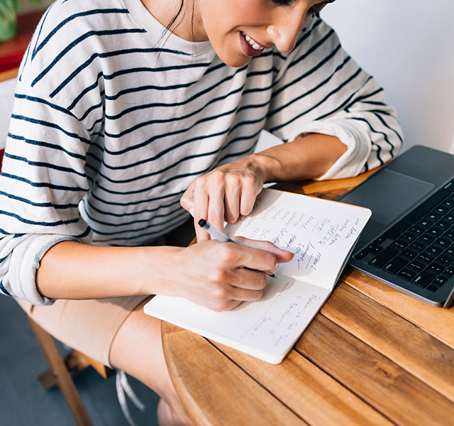 A person in a striped shirt writes notes in a notebook while sitting at a wooden table with a laptop open. handwriting vs typing