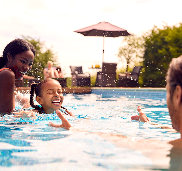 A family of three swimming outside in a pool. The mom is wearing a neon pink two-piece bathing suit and smiling, the daughter has braids in her hair and is smiling and the dad has his arms opened out to his smiling child. dummy mobile image