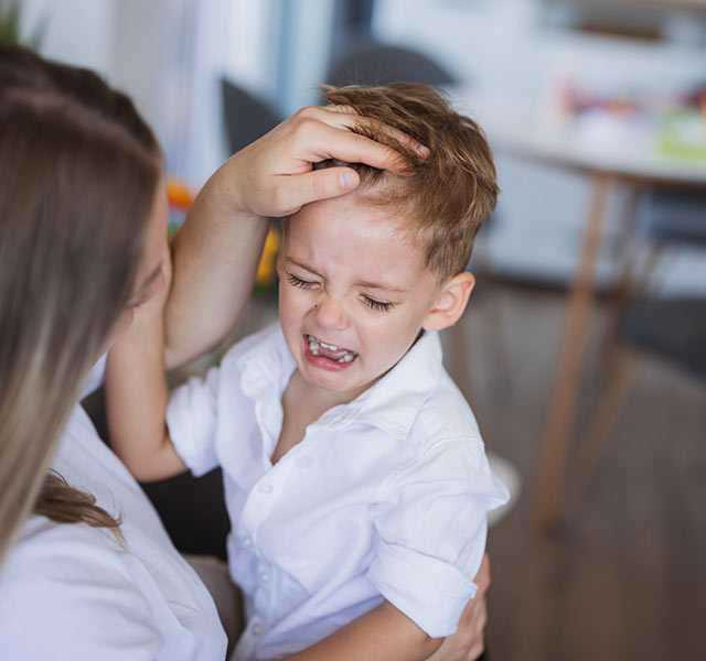 A woman comforting a crying young boy in a home setting. A table and chairs are visible in the background. toddler head bumps
