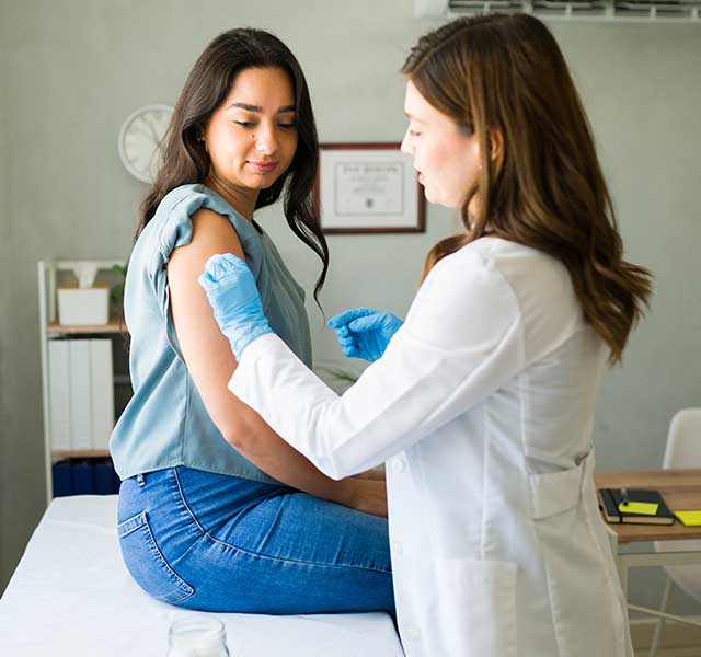 Woman with dark hair sits in doctor's exam room as a female provider prepares to administer a vaccine. flu vaccine 2026