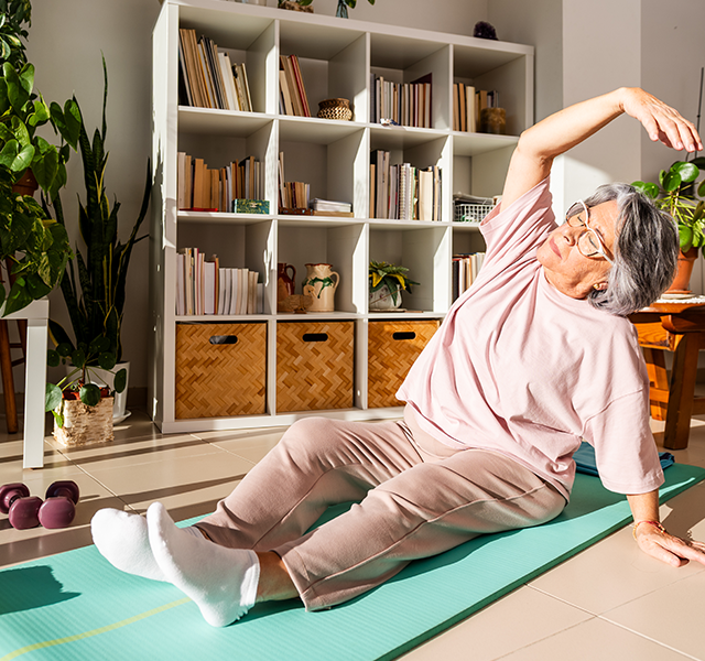 Woman with gray short hair and glasses wearing a nude-colored t-shirt, pants and white socks. She is stretching while sitting on a green yoga mat at home. dummy mobile image