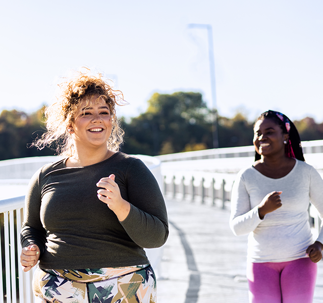 Two friends running outside. One is wearing a black long-sleeved running shirt and multi-colored patterned leggings. The other is wearing a white long-sleeved running shirt and hot pink leggings. Both are smiling. dummy mobile image