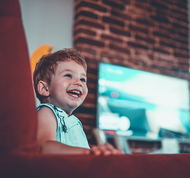 Toddler boy smiling wearing light green overalls. He is sitting on a chair and there is a TV on in the background. dummy mobile image