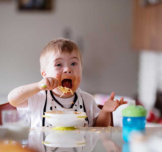 Toddler with Down syndrome happily eating a bowl of pasta. Sauce is on his face and noodles are on the table. eating challenges down syndrome