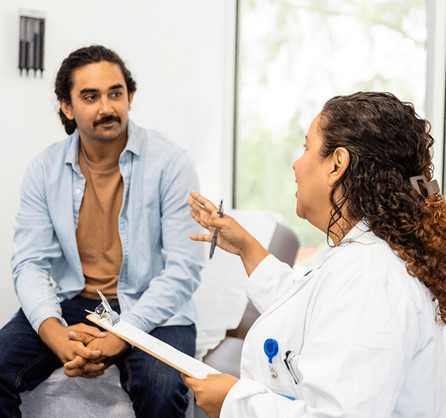 Man with dark hair and a moustache sitting in a doctor's office. He is wearing a light blue button-down shirt with a tan t-shirt underneath, listening to a female doctor talk to him. She has brown curly hair and is wearing a lab coat. dummy mobile image
