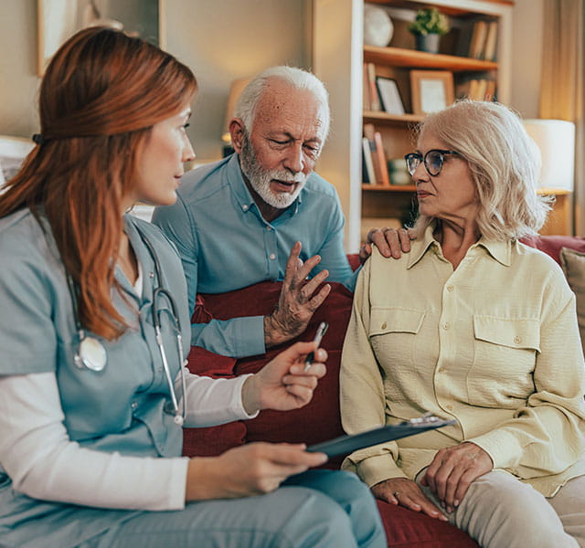 An older couple sits around a couch while talking to a female medical professional. parkinsons and delusions