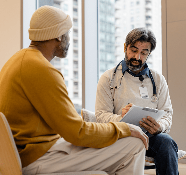 Older man with gray beard, off-white knit hat and marigold-orange sweater talking to older doctor with a black and gray beard wearing a lab coat and stethoscope. He is holding a clip board with a piece of paper on it. dummy mobile image