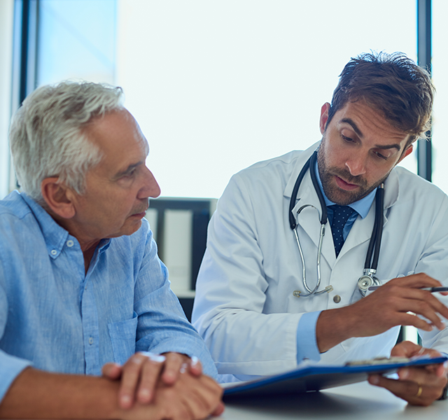 Older man with gray hair wearing a blue button down shirt talking to a male doctor with brown hair wearing a white coat. dummy mobile image