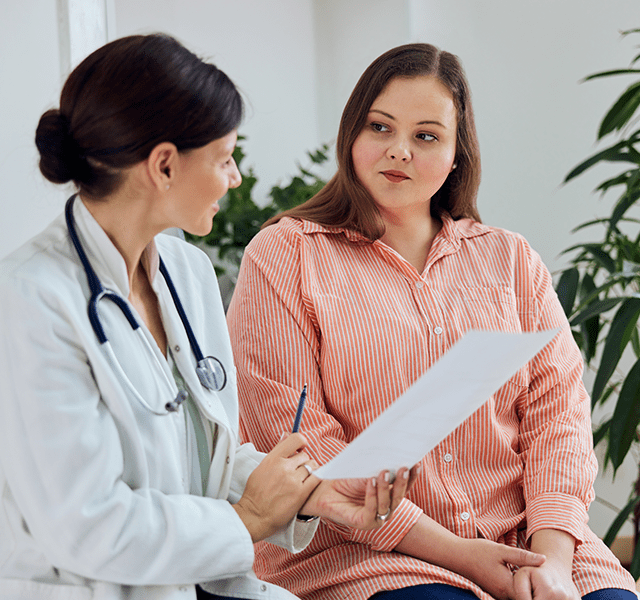 Female doctor with dark brown hair in a low bun wearing a white coat and a stethoscope around her neck. She is talking to a young female patient with long brown hair who is wearing an orange and white vertical striped button-down shirt. dummy mobile image