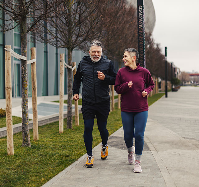 Two people jogging on a sidewalk next to a modern building. They appear to be enjoying the activity together. Trees are planted along the path. The weather is overcast. cognitive health walking speed