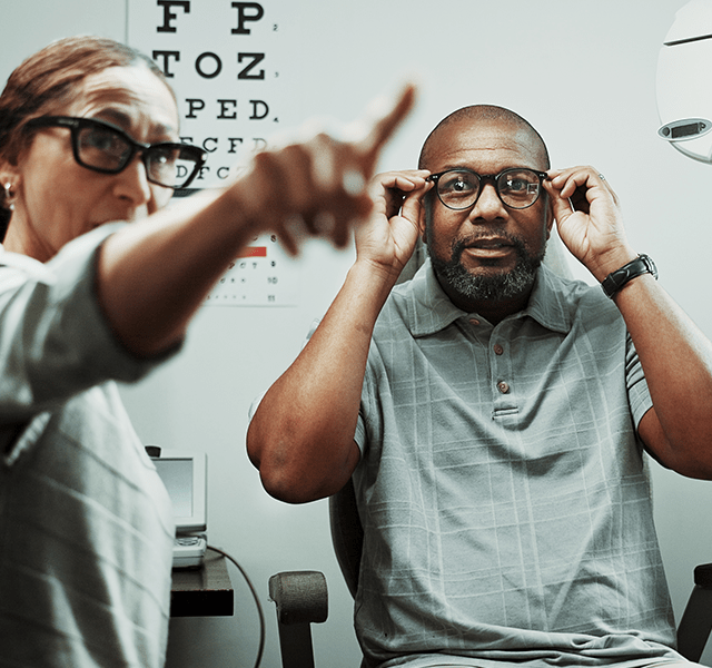 Older man getting an eye exam. He is wearing a gray polo T-shirt and holding glasses up to his eyes while a doctor points at something for him to see. dummy mobile image