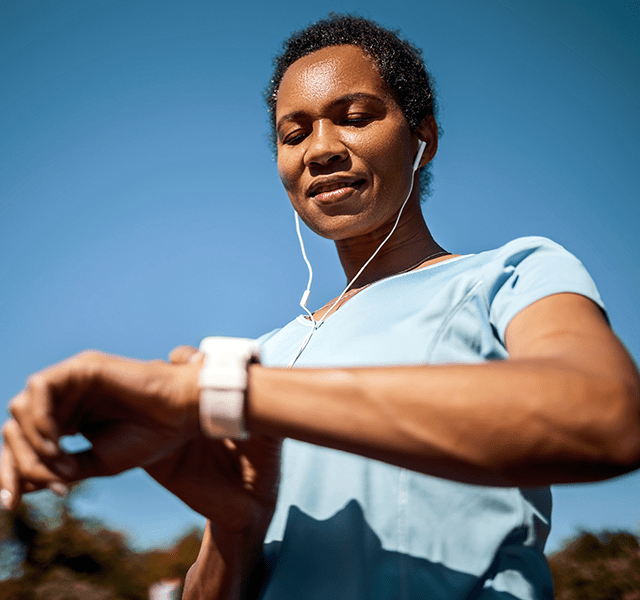 Woman wearing a light blue t-shirt standing outside on a blue-sky day looking at her smart watch. woman checking heart rate on smart watch