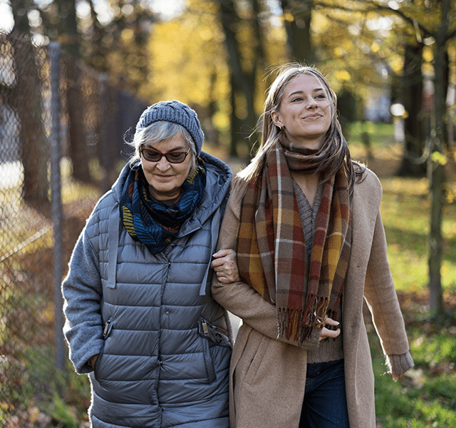 Older woman with gray short hair wearing a blue hat, gray puffer coat, teal scarf and sunglasses. She is walking outside with a younger woman who has blonde hair and is wearing a camel coat and a multi-colored plaid scarf. dummy mobile image