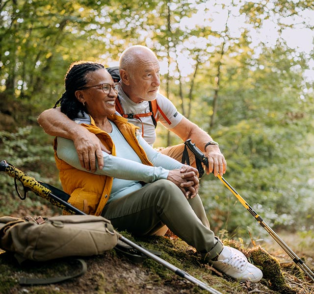A man with short white hair and a backpack is sitting on a hillside, arm around a woman with long braided hair and glasses. Both are smiling and holding hiking poles, surrounded by lush green trees and sunlight. preventing osteoporosis