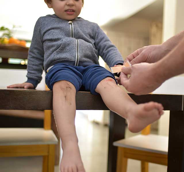 A little boy sits on the counter while his father puts a bandage on his scraped knee. treating minor wounds