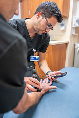 Resident physician performs a hand examination while reviewing clinical information on a mobile device at Henry Ford Health Seville Family Medicine clinic.
