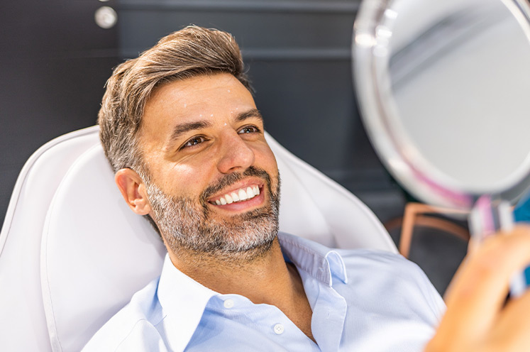 Bearded man sitting in recliner exam room chair holding handheld mirror and looking at his face while smiling